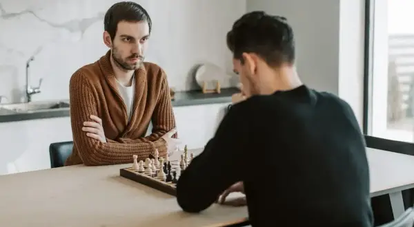 Dwóch mężczyzn gra w szachy. Jeden z nich, w brązowym swetrze, zamyślony patrzy na planszę, gotowy na gambit.