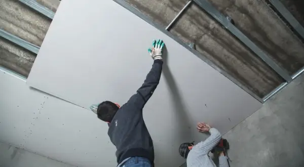Deux ouvriers s'affairent à habiller un plafond abîmé avec des plaques de plâtre, fixées sur une structure métallique.