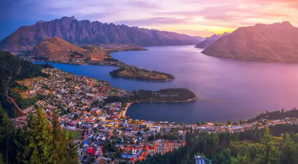 Vacker solnedgång över Queenstown, Nya Zeeland. Staden ligger vid en sjö, omgiven av berg och skog.