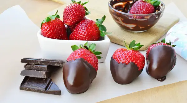 Fragole fresche e succose, alcune immerse in cioccolato fuso, altre pronte per esserlo, accanto a tavolette di cioccolato fondente.