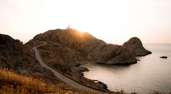 Chemin sinueux menant à un phare sur une colline rocheuse, une porte de pierre naturelle s'ouvrant sur la mer au coucher du soleil.