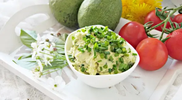 Pyszna pasta z awokado, jajkiem i szczypiorkiem, podana z pomidorkami i kwiatami na białej tacy.
