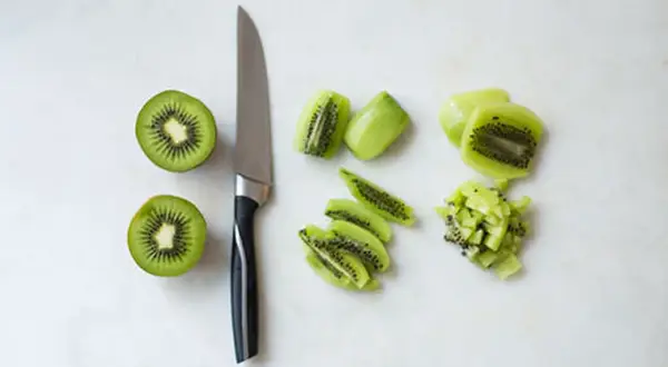 Ein Messer liegt neben zwei halbierten Kiwis. Daneben sind die Früchte in Scheiben und Würfel geschnitten, bereit zum kiwi schneiden.