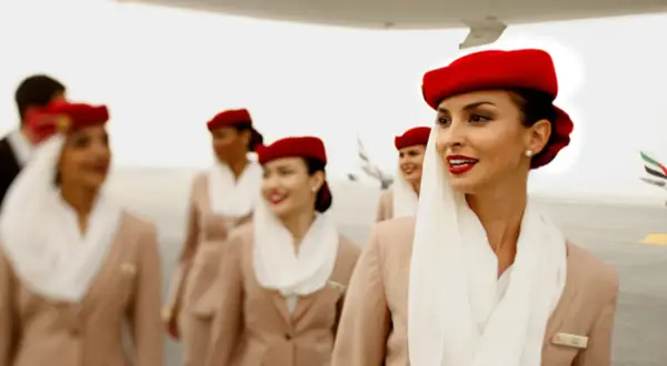 Uśmiechnięte stewardessy Emirates w czerwonych beretach i beżowych uniformach idą po płycie lotniska.