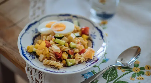En färgglad pastasallad med ägg, grönsaker och ost, perfekt som pastasallad barn. En sked och ett glas vatten står bredvid.