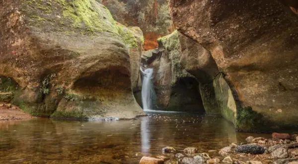 Cascada en el gorg de la Mola, rodeada de rocas cubiertas de musgo y vegetación. El agua clara refleja el entorno natural.