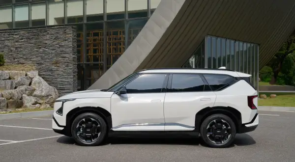 Nowy, biały suv Kia EV9 stoi zaparkowany przed nowoczesnym budynkiem z kamienną fasadą i dużymi oknami.