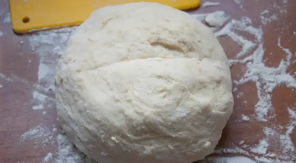 Surowe ciasto na pizzę na drewnianym blacie, posypane mąką. Obok żółta łopatka.
