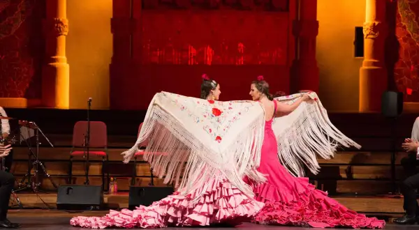 Dos bailarinas de flamenco, con vestidos rosas y mantones bordados, despliegan su arte. El escenario tiene un aire de tener aje.