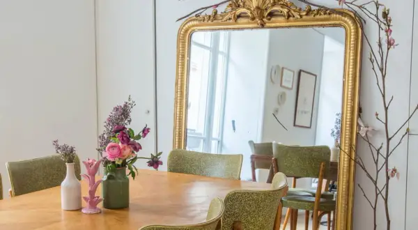 Un grand miroir au cadre doré orné de fleurs et de branches. Une table en bois est dressée avec des vases fleuris, prête à repeindre son cadre doré.
