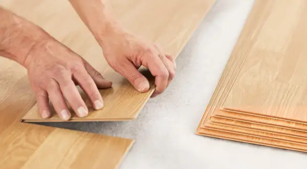 Hands installing laminate flooring over a subfloor. A stack of laminate planks is visible on the right. This shows how you can put laminate flooring over vinyl.