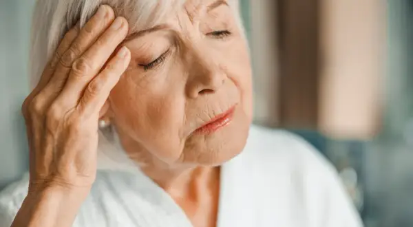Starsza kobieta z ręką na skroni, zamyślona. Może to być jeden z objawów choroby Alzheimera.