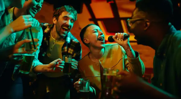 Freunde singen lautstark bei Karaoke mit, halten Getränke und haben sichtlich Spaß. Perfekt für lieder zum mitsingen!