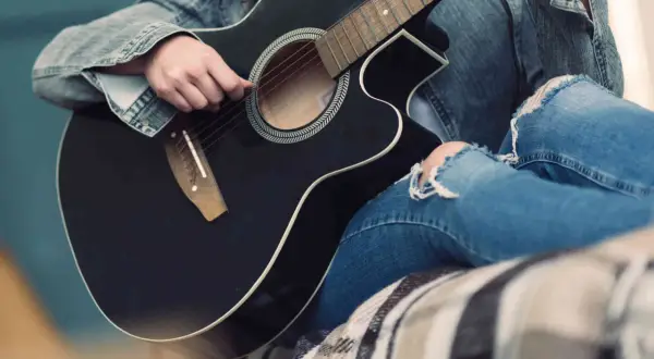 Osoba w dżinsowej kurtce i podartych jeansach gra na czarnej gitarze. Słychać delikatne dźwięki, jakby tworzyła własną melodię.