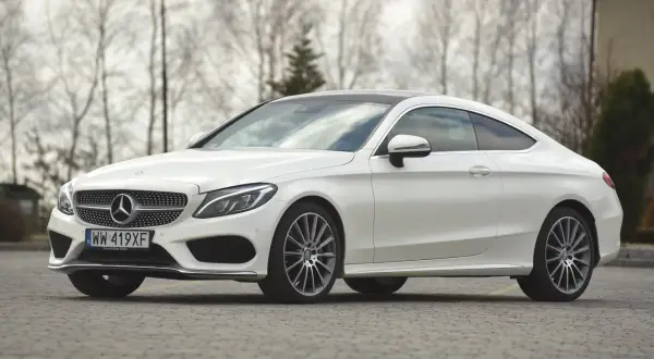 Biały Mercedes C klasa coupe z czarnym dachem, zaparkowany na brukowanej nawierzchni, z drzewami w tle.