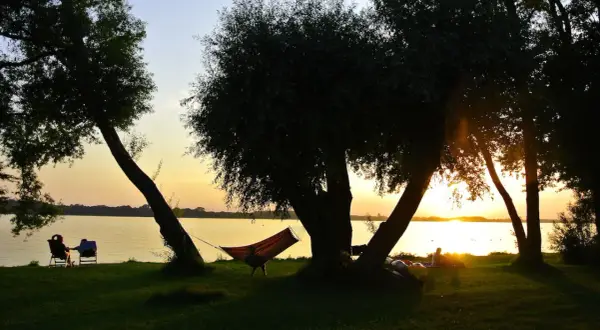 Sonnenuntergang am See mit Hängematte und Liegestühlen im Schatten von Bäumen.