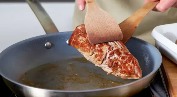 Ein saftiges Schweinefilet braten in der Pfanne. Mit zwei Holzspateln wird das Fleisch gewendet, um es von allen Seiten perfekt zu garen.