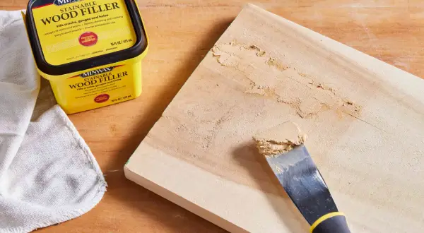 Un pot de mastic à bois Minwax est à côté d'une planche de bois où un couteau à mastic applique le produit pour reboucher bois.