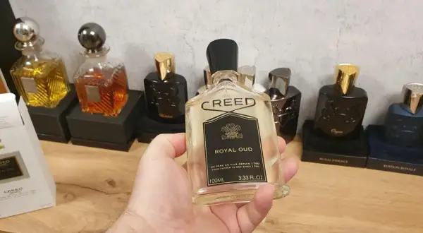 Kolekcja perfum Creed, w tym Creed Royal Oud, prezentowana na drewnianym blacie. W dłoni trzymana jest butelka Creed Royal Oud.