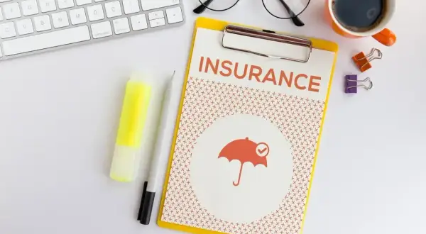 Planujesz ubezpieczenie zdrowotne bez pracy? Na biurku dokument "INSURANCE" z parasolką, klawiatura, okulary i kawa.
