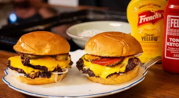 Två saftiga smashed burgers med smält ost, pickles och lök. Bredvid står flaskor med senap och ketchup.