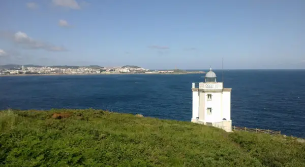 El faro de Mera se alza blanco sobre un promontorio verde, vigilando la costa y el mar azul.