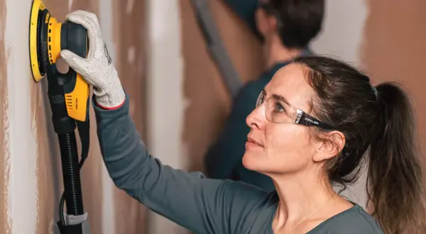 Donna con occhiali protettivi usa una levigatrice per imparare come levigare un muro.