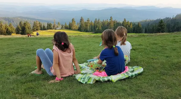 Trzy dziewczynki siedzą na kocu na trawie, podziwiając widok z hali Rysianka.