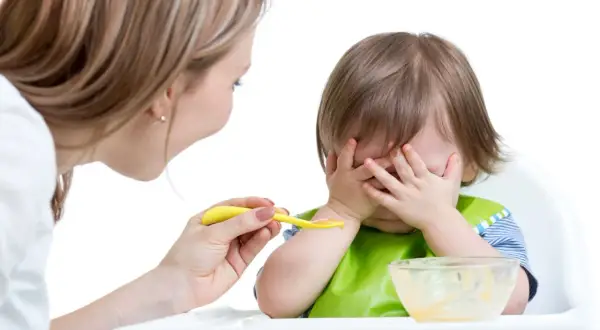 Mama próbuje nakarmić malucha, ale dziecko nie chce jeść, zakrywając buzię rączkami.