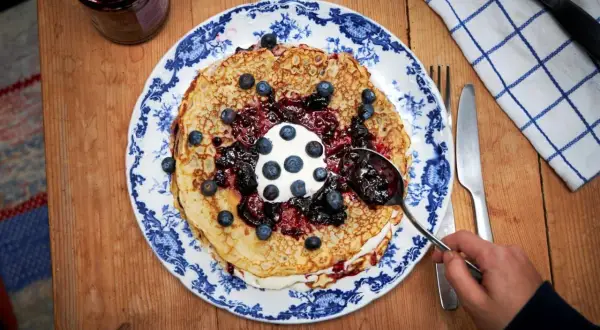 En hög med pannkakor toppas med grädde, blåbär och en generös klick drottningsylt. Perfekt för ett mysigt fika.