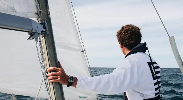 Ein junger Mann mit lockigem Haar blickt aufs Meer, während er die Hand am Mast hält. Er ist bereit, die Segel zu setzen und in See zu stechen.