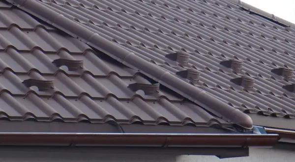 Brązowy dach z dachówki ceramicznej, widoczne elementy systemu rynnowego i wentylacyjnego. Jak zrobić zakończenie gąsiora?