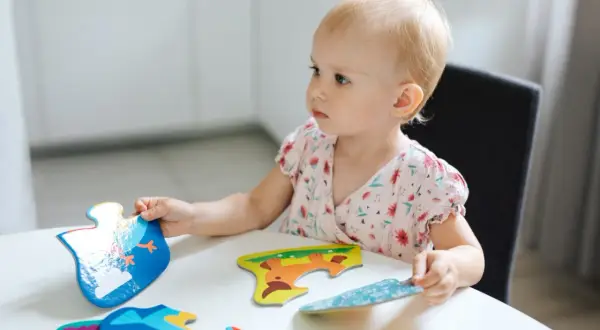 Mała dziewczynka z blond włosami skupiona na układaniu kolorowych puzzli z zwierzątkami.