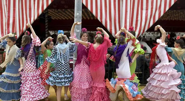 Niñas con trajes de volantes y lunares, aprendiendo a bailar sevillanas con arte bajo una carpa a rayas.