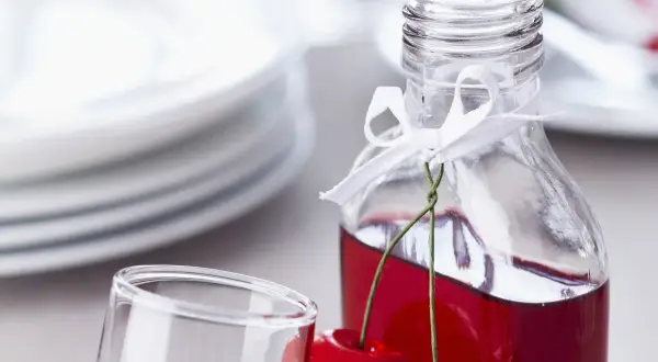 Elegancka butelka z wiśniowym alkoholem, ozdobiona wstążką i świeżymi wiśniami, obok kieliszka i stosu talerzy.