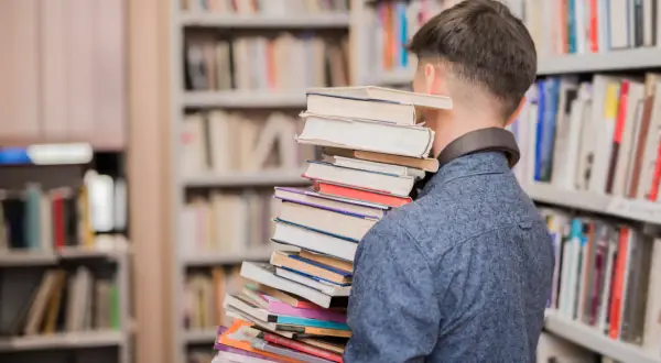 Młody mężczyzna w bibliotece dźwiga stos książek, symbolizując przejście od statusu ucznia do statusu studenta.