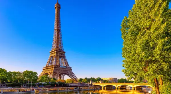 Eiffeltornet i Paris, med sina tre våningar, speglar sig i Seine. En grönskande trädtopp skymtar till höger.