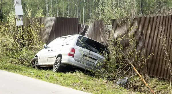 Srebrny minivan wjechał w drewniane ogrodzenie, powodując jego uszkodzenie. Pojazd jest częściowo wciśnięty w płot.