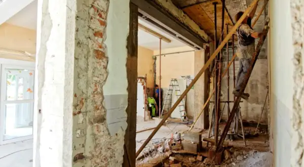 Remont wnętrza, pracownik na drabinie przy ścianie nośnej. Widać cegły i gruz.