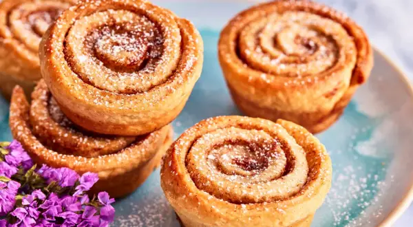 Zimtschnecken Muffins, bestäubt mit Puderzucker, liegen auf einem blauen Teller neben lila Blumen.