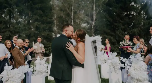 Para młoda całująca się podczas ceremonii ślubnej na zewnątrz, otoczona gośćmi i dekoracjami.