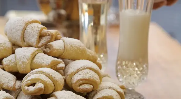 Stos rogalików krucho-drożdżowych z 1 kg mąki, posypanych cukrem pudrem, obok kieliszków z napojem.