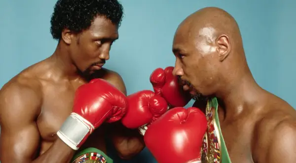 Two boxers, one with curly hair, the other bald, face off in a tense stare-down, red gloves raised, hinting at the legendary Marvin Hagler war.