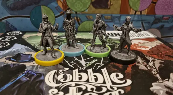 Cztery figurki postaci z gry planszowej "Cobble & Fog" na tle planszy. Atmosfera unmatched bruk i mgła.