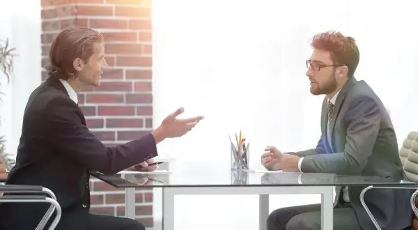 Dwóch mężczyzn w garniturach siedzi przy biurku. Jeden gestykuluje, jakby omawiał, czy na rozmowie kwalifikacyjnej pytać o zarobki.
