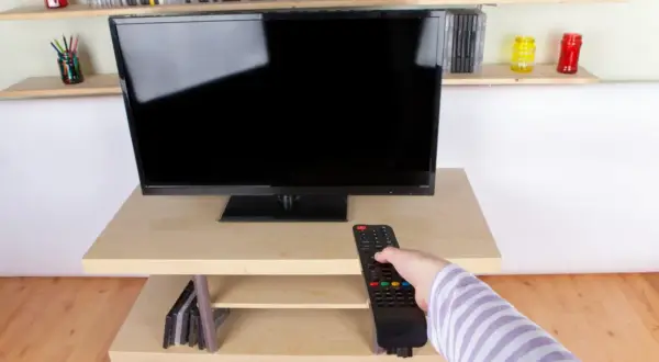 Ręka z pilotem sięga do telewizora naprzeciwko okna. Na półkach stoją płyty i kolorowe słoiki.