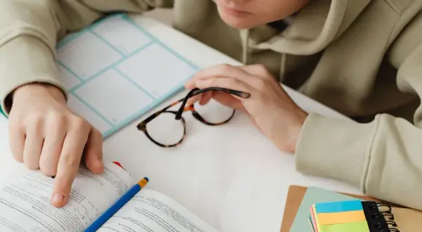 Osoba w bluzie z kapturem przegląda książkę, obok leżą okulary i kolorowe karteczki.