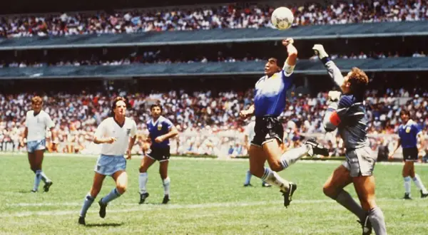 Diego Maradona, w słynnym meczu, używa "ręka boga", by zdobyć bramkę. Piłkarze w białych strojach patrzą z niedowierzaniem.