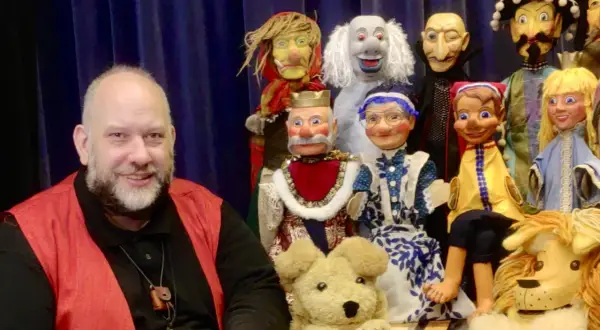 Ein Mann lächelt neben einer bunten Truppe von Handpuppen, die für den Puppenpavillon Bensberg bereitstehen.