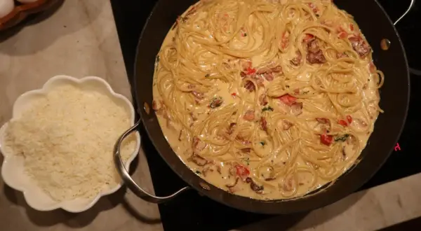 En stor panna med krämig pasta med baconsås. Bredvid en skål med riven ost.